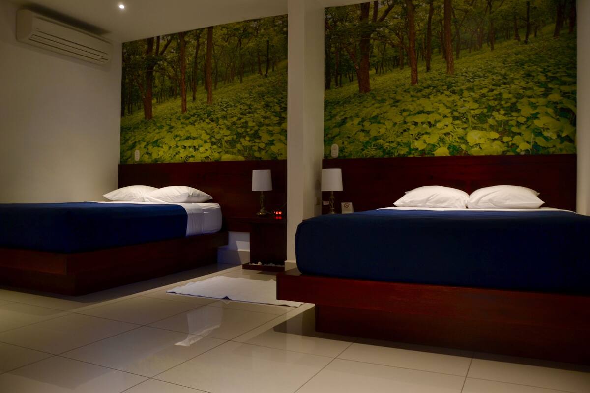 A hotel room with two beds, a large forest mural on the wall, a bedside table with a lamp, and a modern design.
