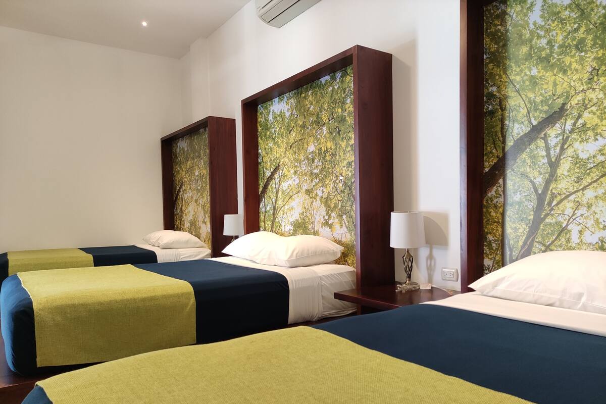 A hotel room with three beds, each with a different colored bedspread. There are two wall-mounted lamps on wooden nightstands.