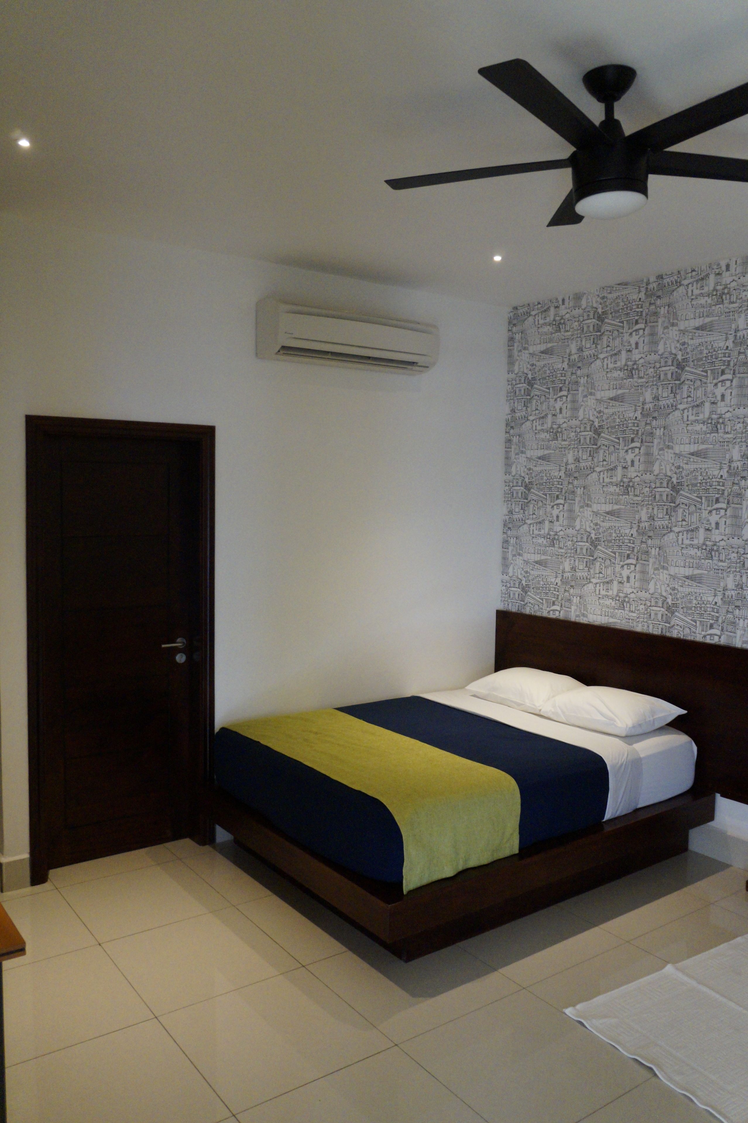 A bedroom with a bed, a ceiling fan, an air conditioner, a patterned wallpaper, and a door.
