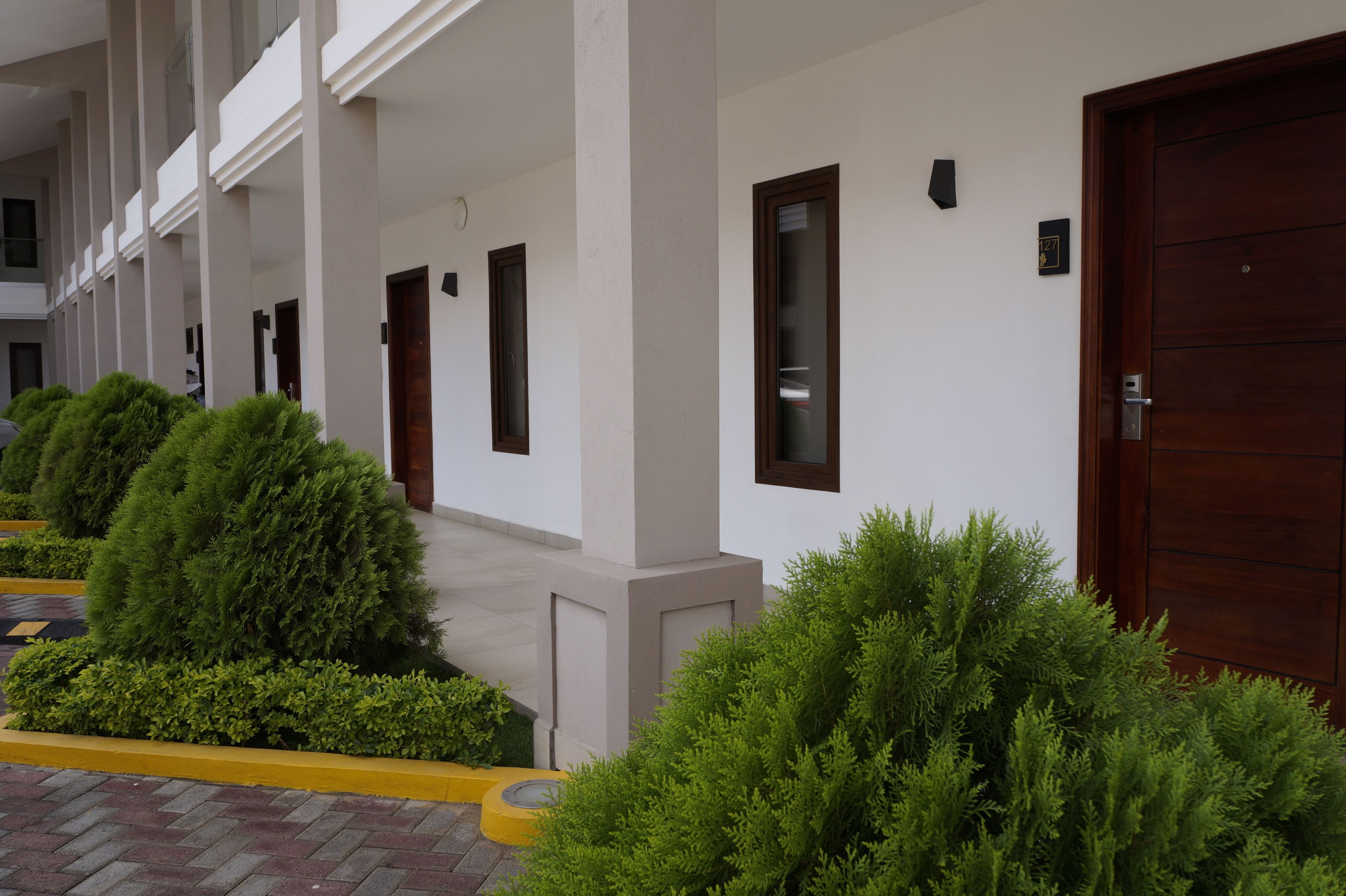 A hotel corridor with multiple doors, a covered walkway, and neatly trimmed bushes.