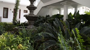 A fountain surrounded by lush greenery in a courtyard with white columns and a multi-story building in the background. - Hotel Agualcas (Managua)