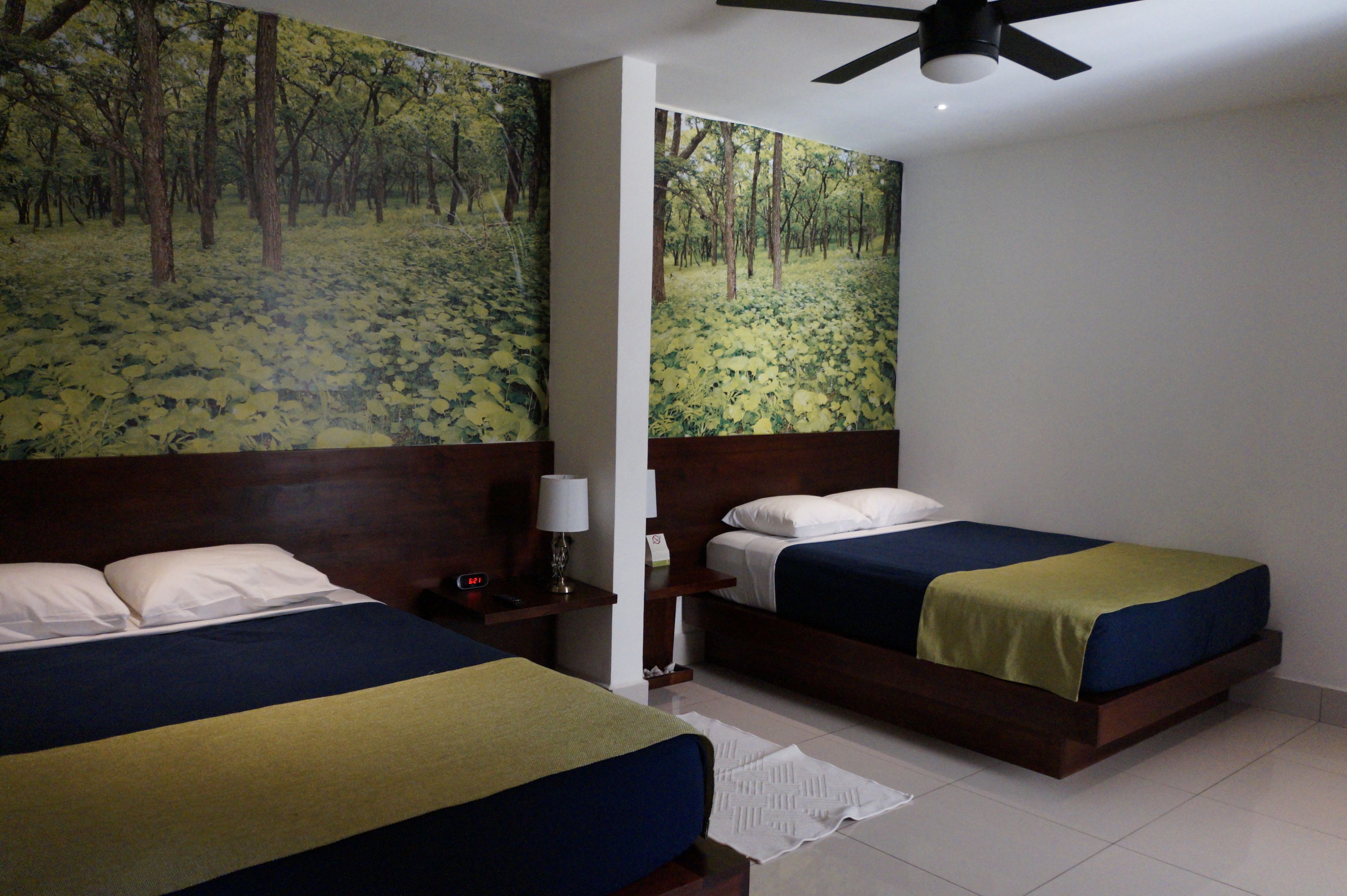 Habitación de hotel con dos camas, un ventilador de techo y un gran mural de un bosque en la pared.