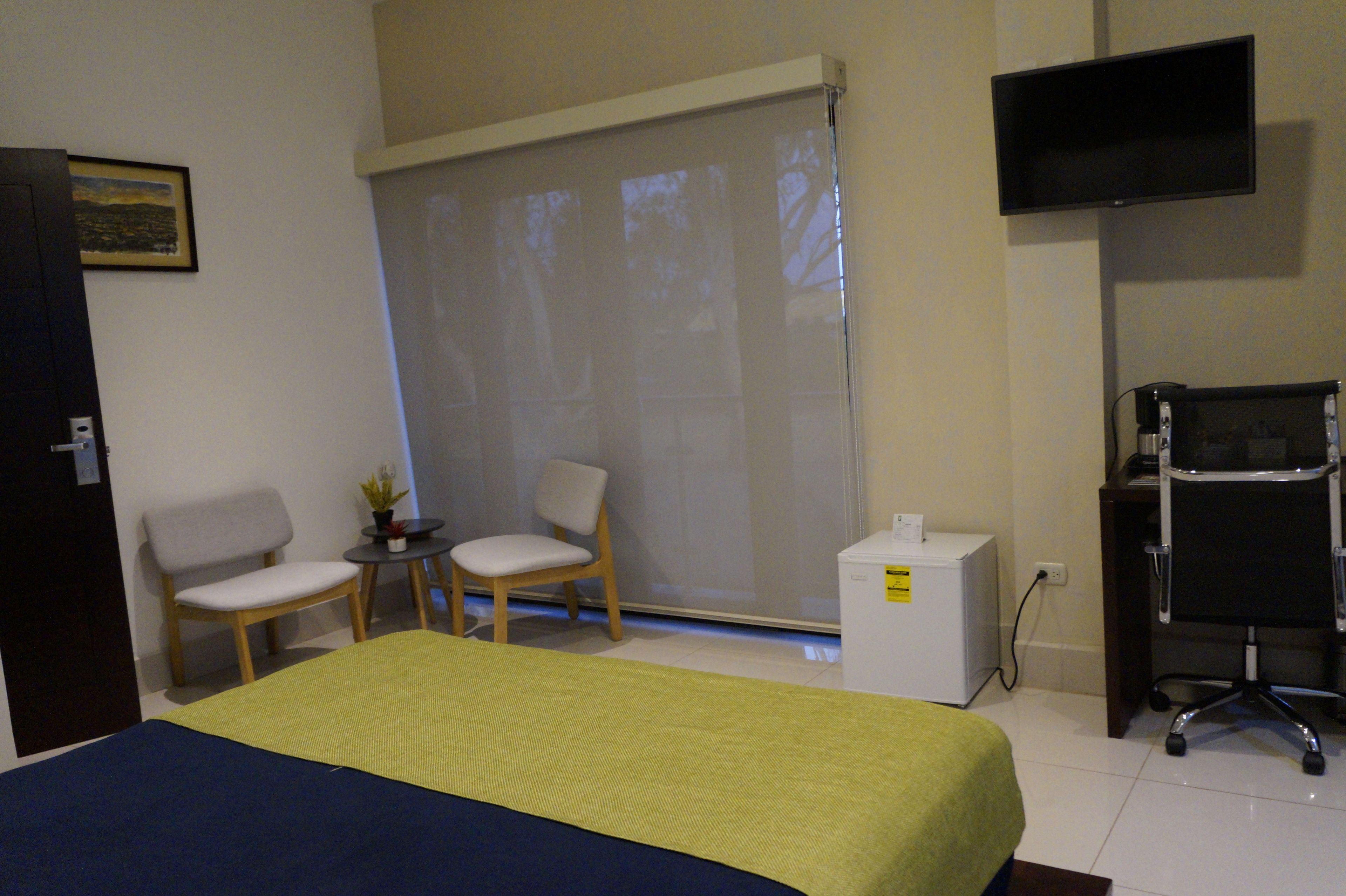 A hotel room with a bed, two chairs, a small table with a plant, a desk with a computer, and a TV on the wall.