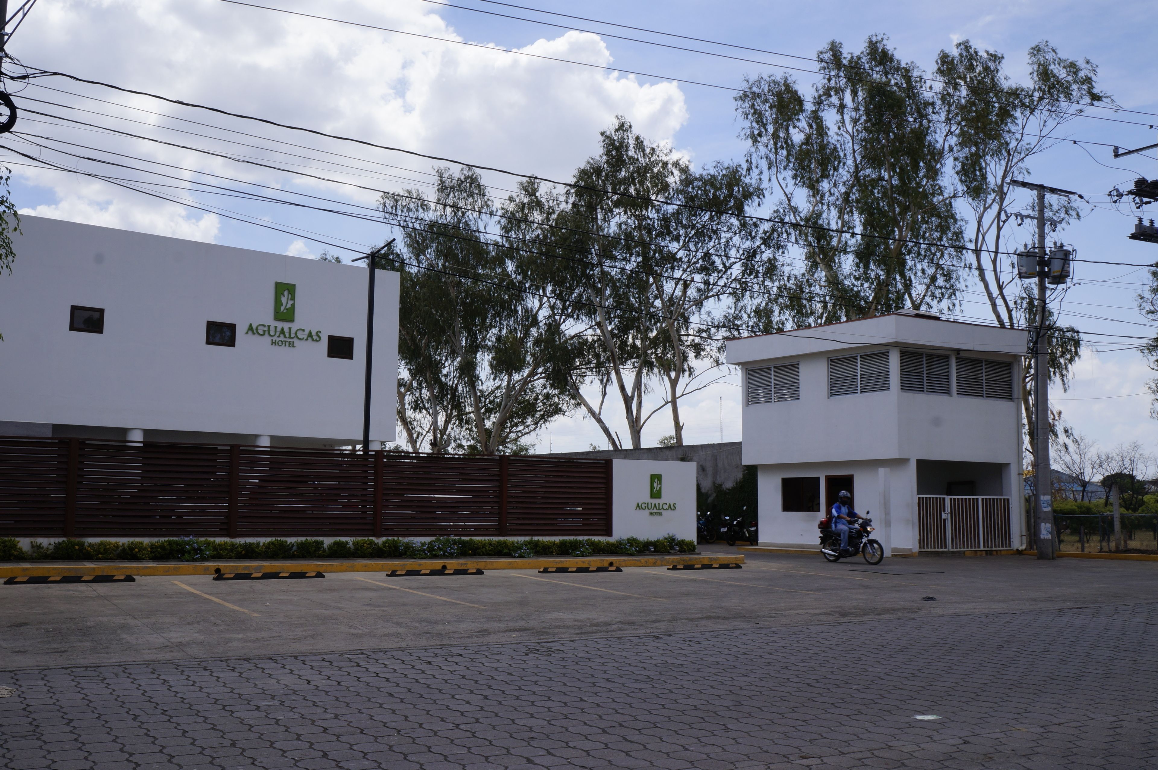 Un hotel moderno con fachada blanca, entrada protegida por una reja y zona de estacionamiento pavimentada.