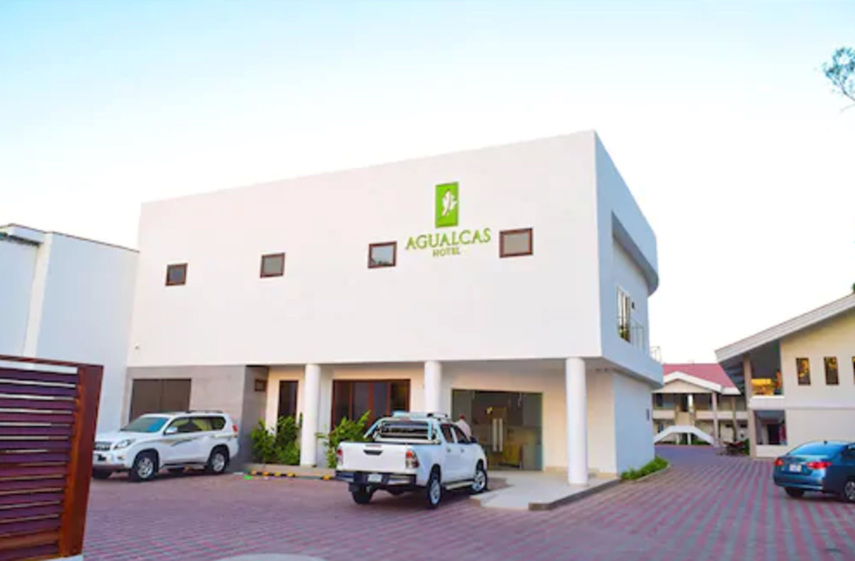 A modern hotel building with a white facade and a green logo. There are multiple vehicles parked in front, including a white SUV and a white pickup truck.