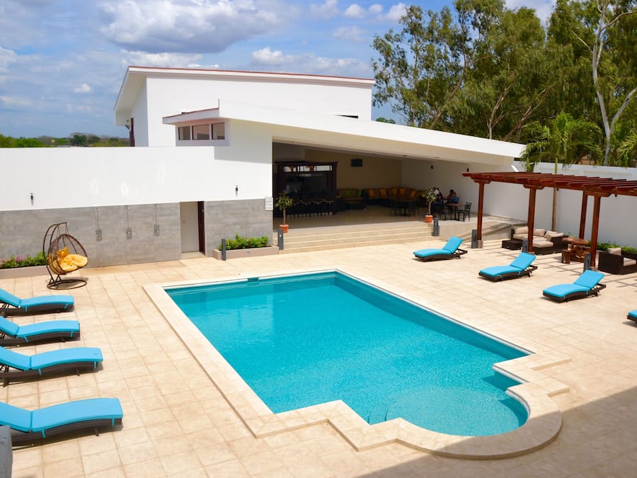 Un área de piscina moderna con una piscina rectangular, sillas de descanso azules y una zona de estar cubierta.