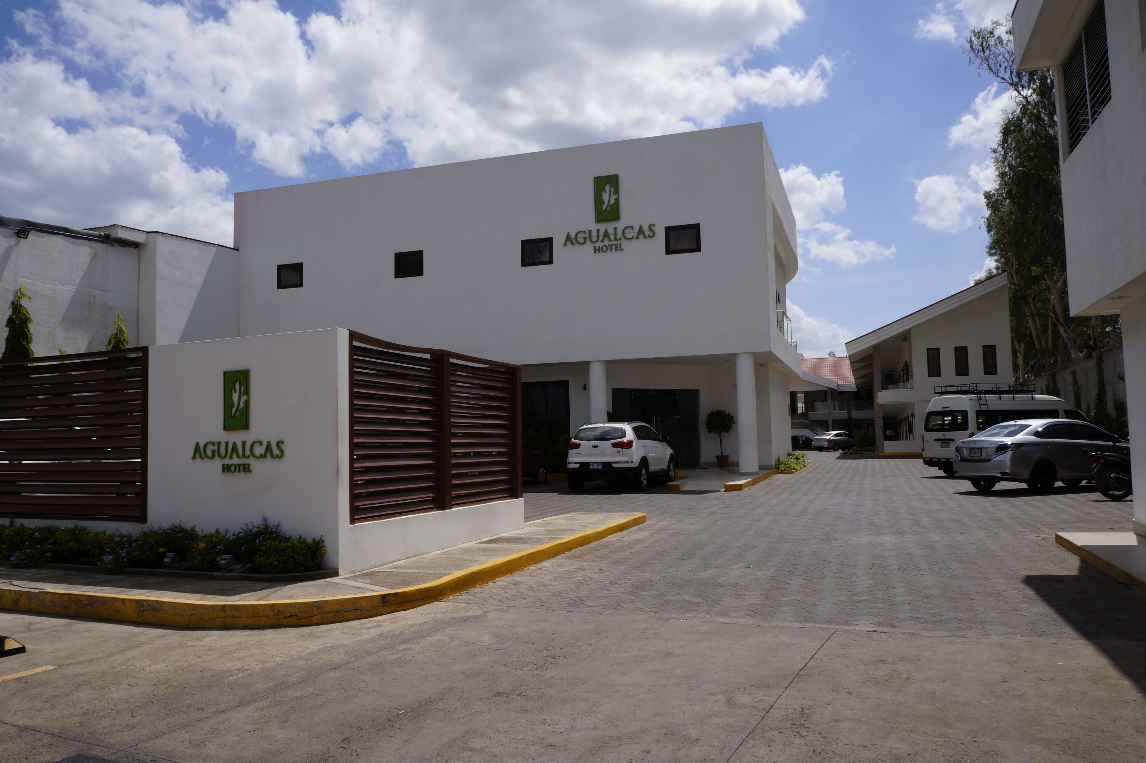 A modern hotel with a flat roof, a central entrance, and a parking area with several cars.