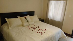 Una cama bien hecha con ropa de cama blanca y una sola rosa sobre la almohada.