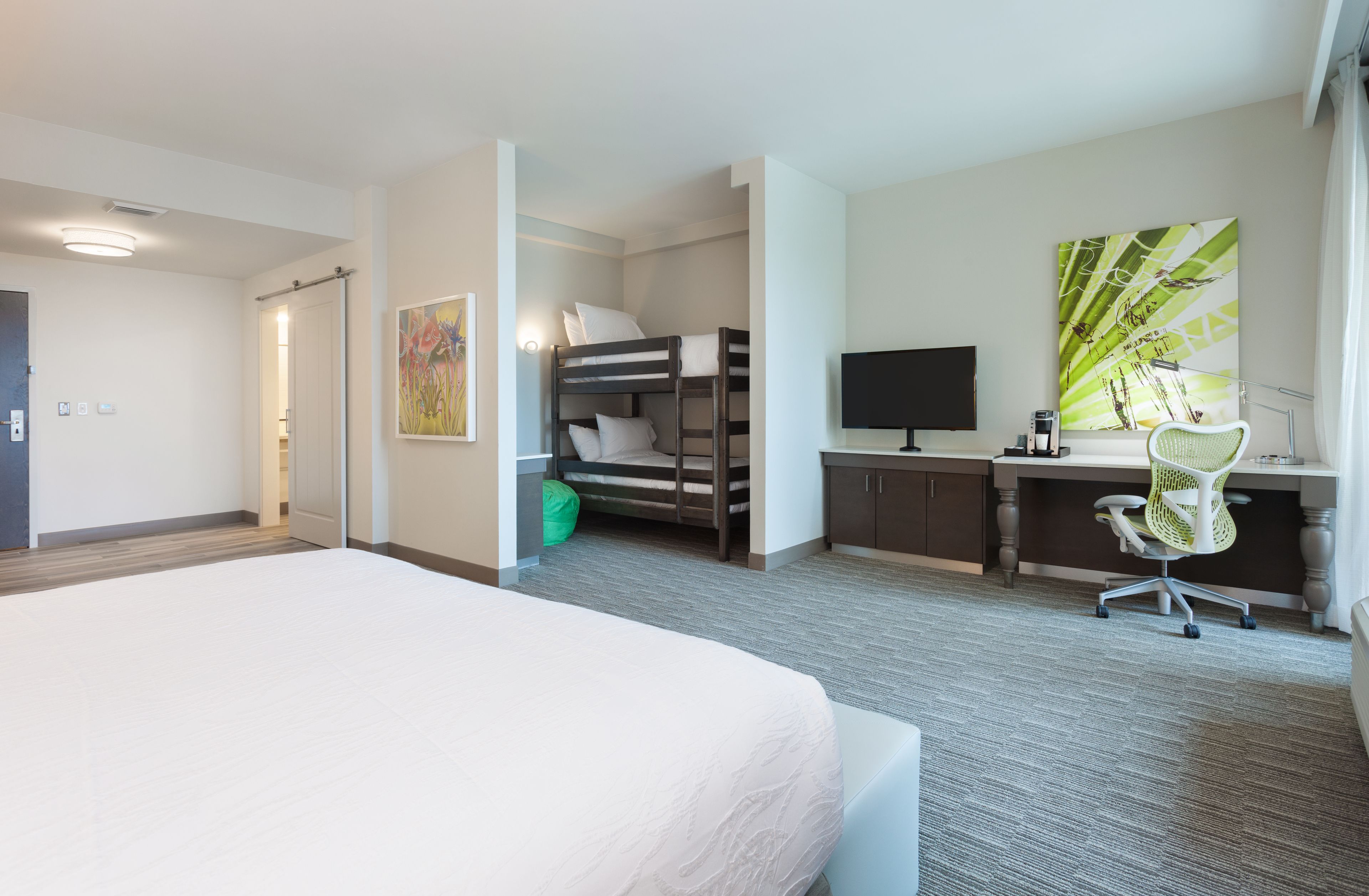 A hotel room with a large bed, a desk with a computer, a chair, a bunk bed, and a TV.