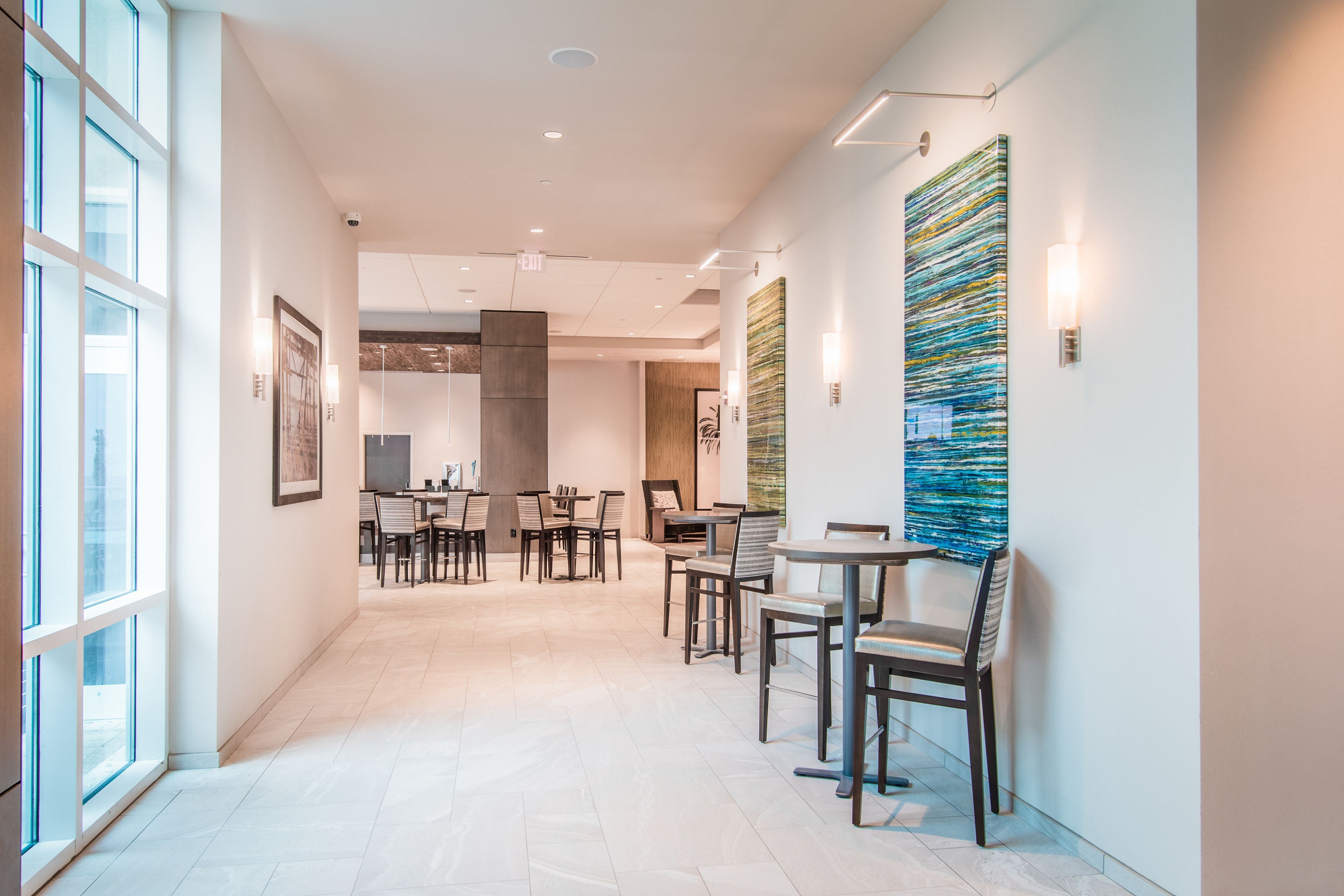 Un hall d’hôtel moderne avec un long couloir, un espace détente doté de tables et de chaises, et de grandes fenêtres sur un côté.