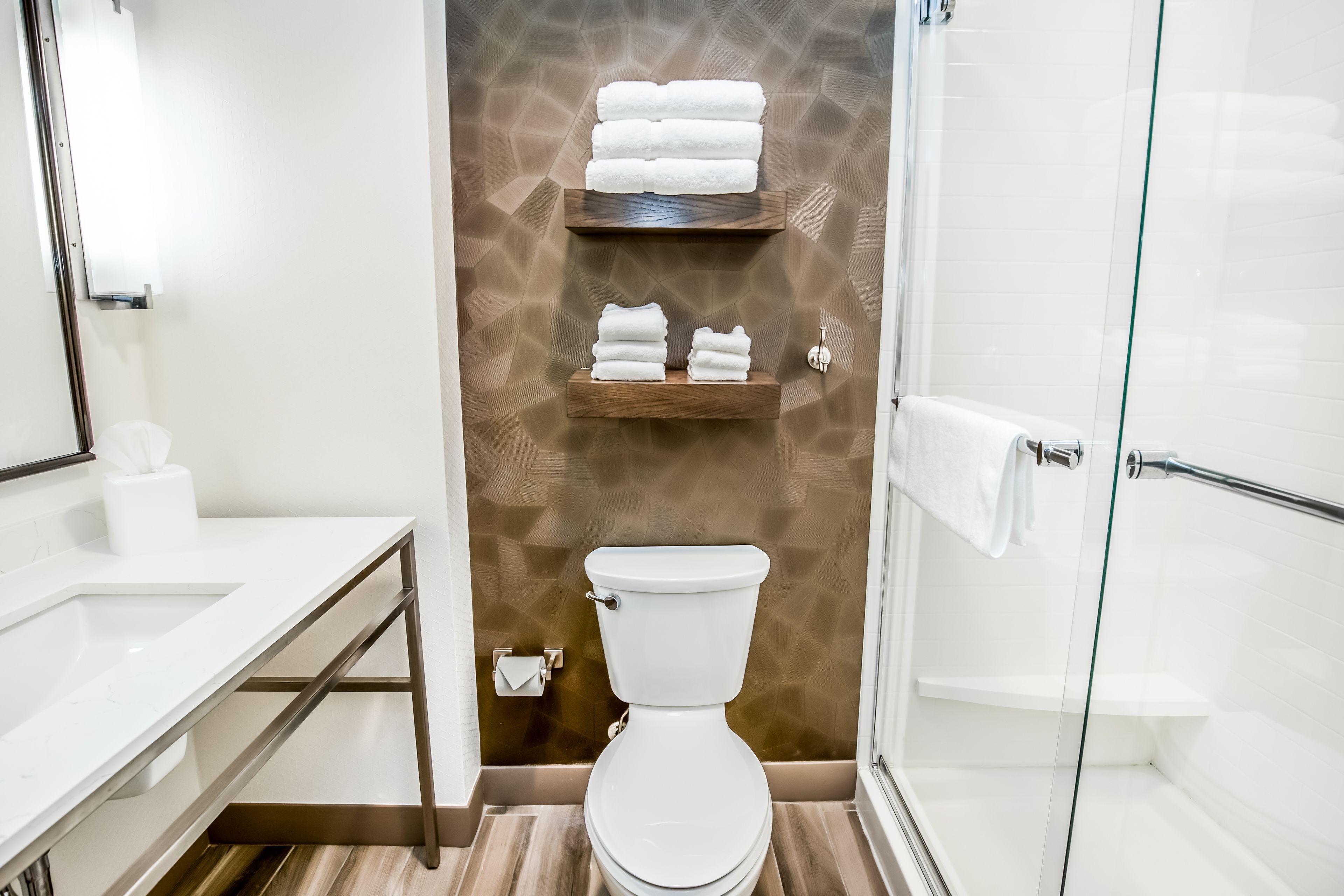 Une salle de bain moderne équipée de toilettes, d’un lavabo, d’une douche en verre et d’étagères en bois où sont posées des serviettes.