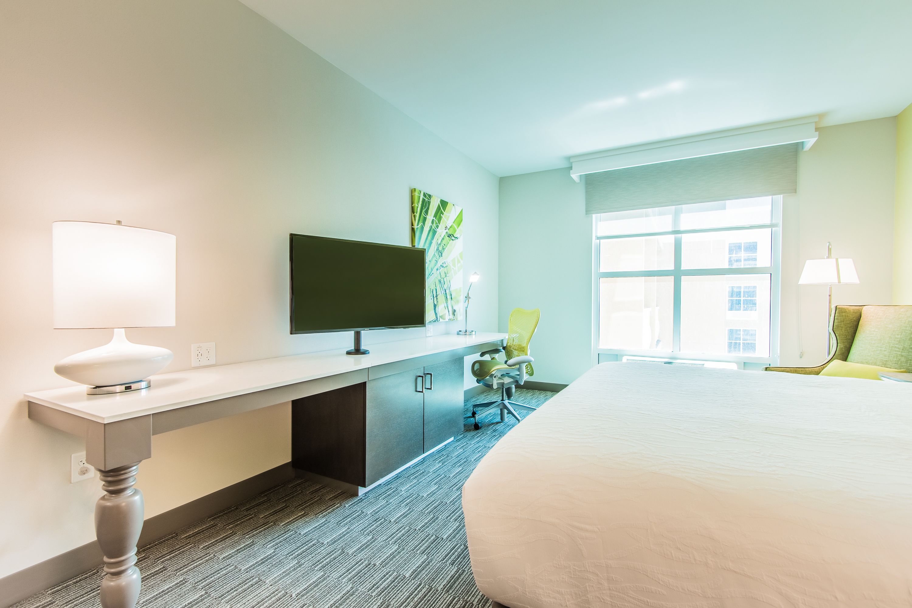 A hotel room with a bed, a desk with a TV, a chair, and a window.