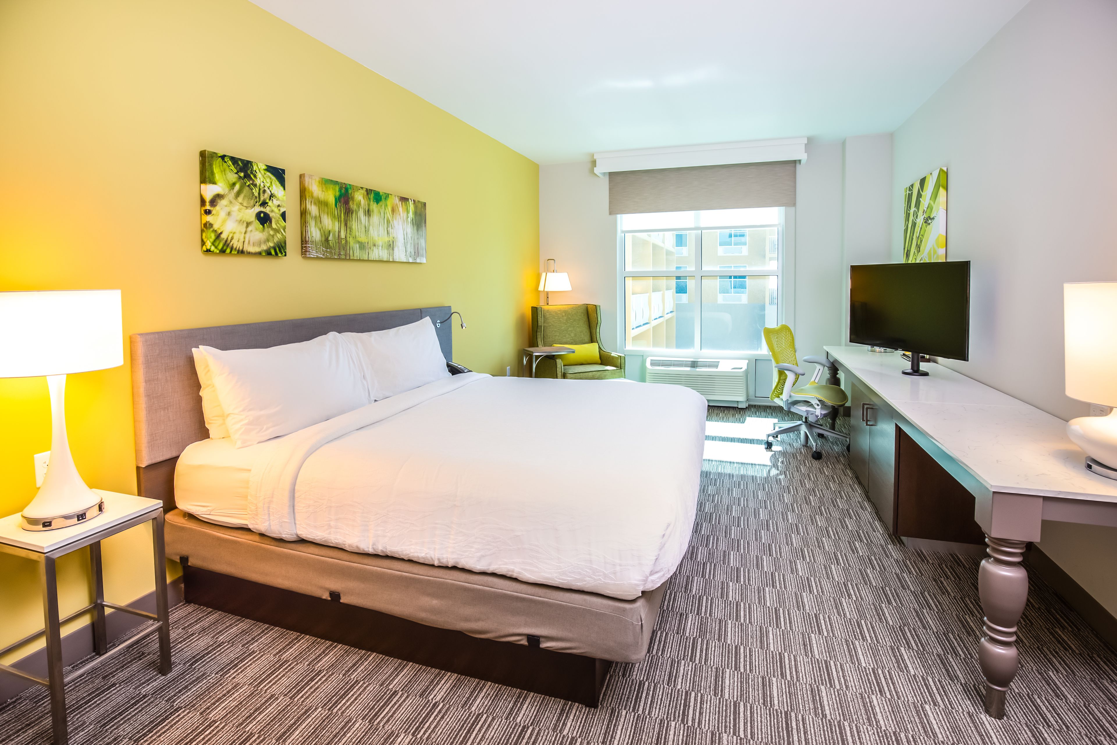 A hotel room with a bed, a desk, a chair, a TV, and artwork on the walls.