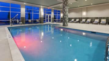An indoor swimming pool with clear blue water, surrounded by lounge chairs and a large window with a view of the evening sky.