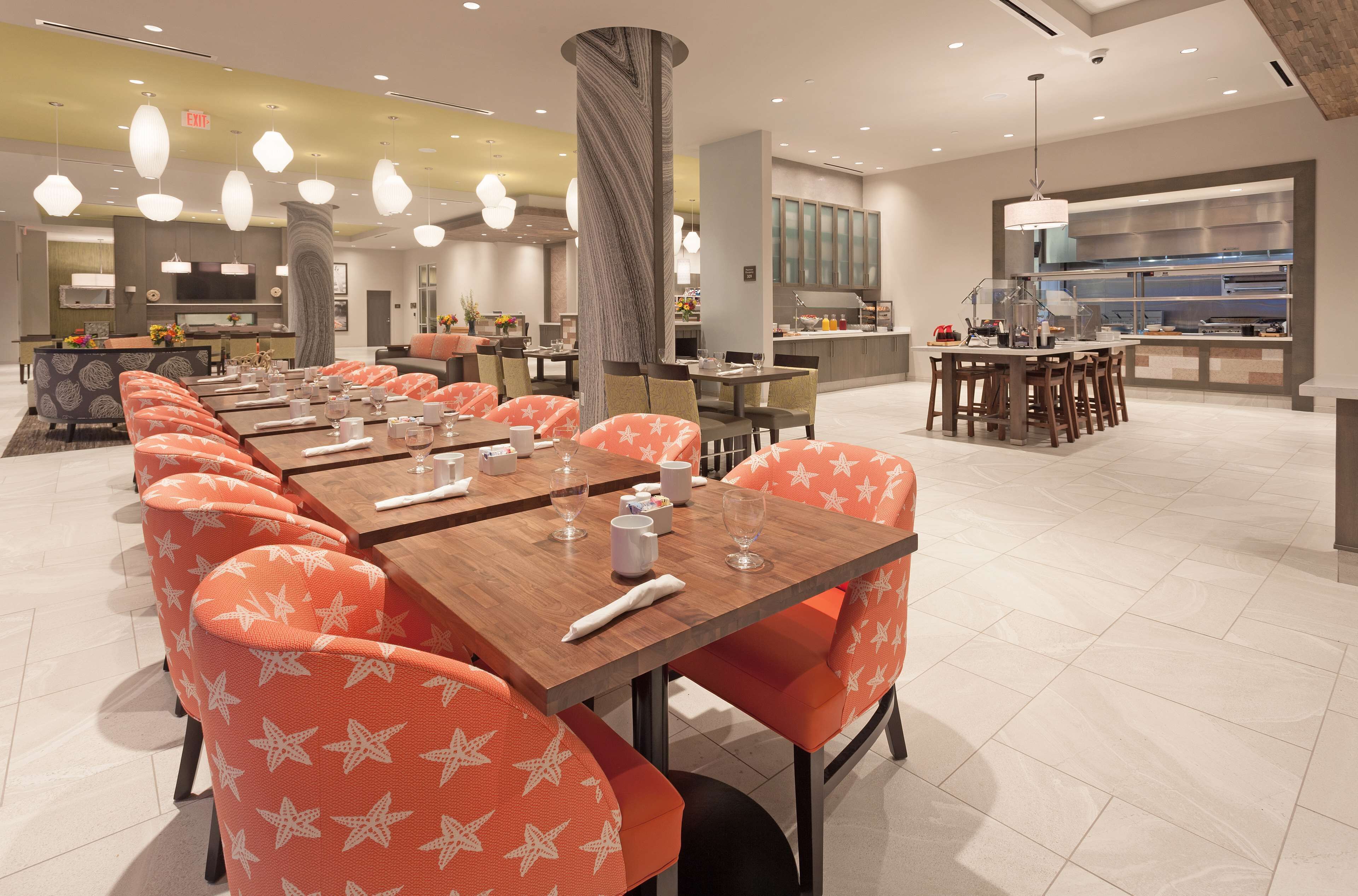 Un comedor moderno con mesas largas y sillas anaranjadas con diseños de estrellas blancas, un pilar central y una zona de cocina al fondo.