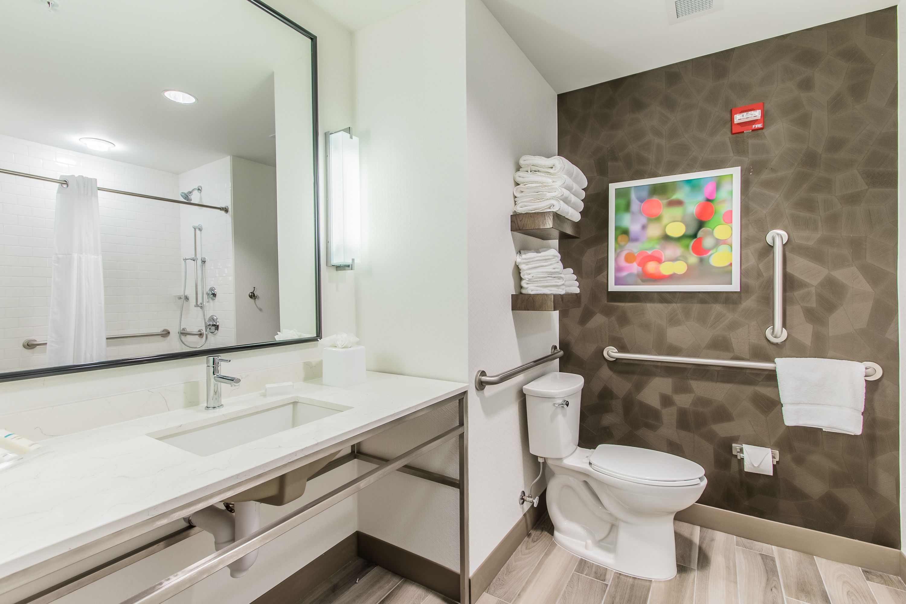 A modern bathroom with a white sink, mirror, and toilet. There are towels stacked on a shelf and a handrail for accessibility.
