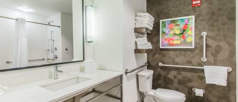 A modern bathroom with a white sink, mirror, and toilet. There are towels stacked on a shelf and a handrail for accessibility.