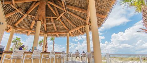 Un espacio de estar al aire libre con techo de paja y vigas de madera, con sillas de metal blanco y vistas al cielo y palmeras.