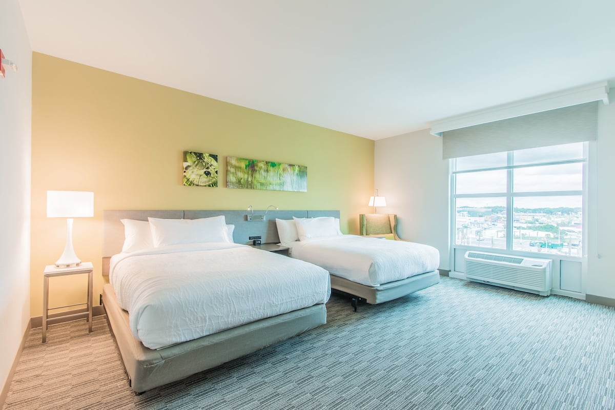 A hotel room with two beds, a large window with a city view, and a wall-mounted air conditioner.