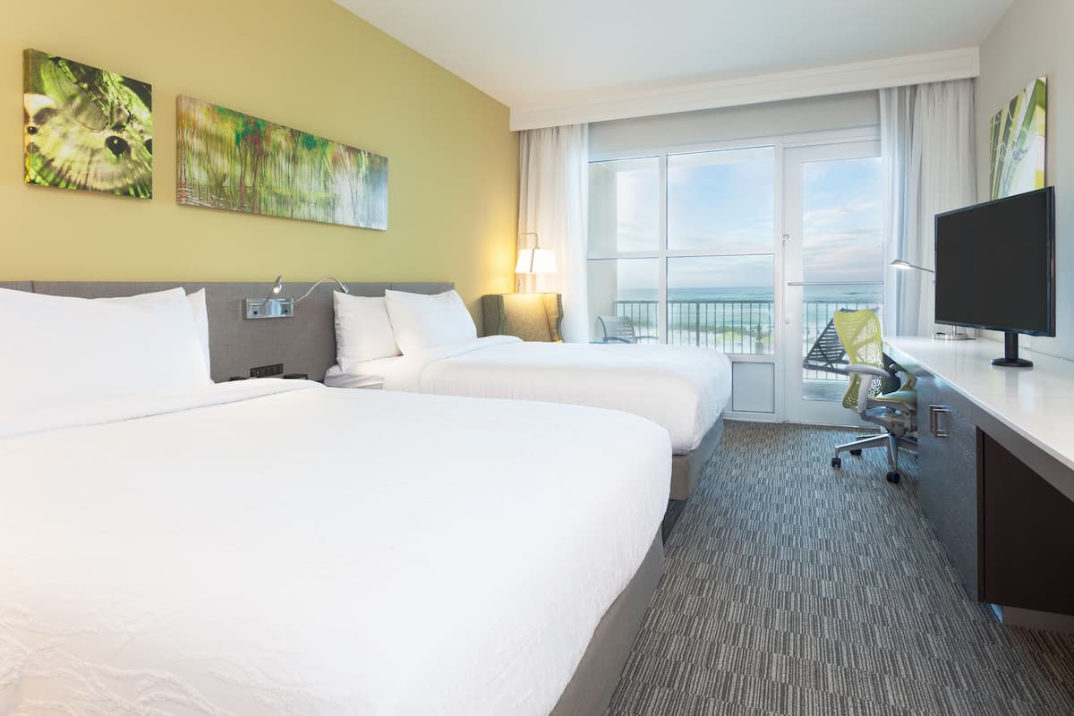 A hotel room with two beds, a desk, a TV, and a balcony with a view.