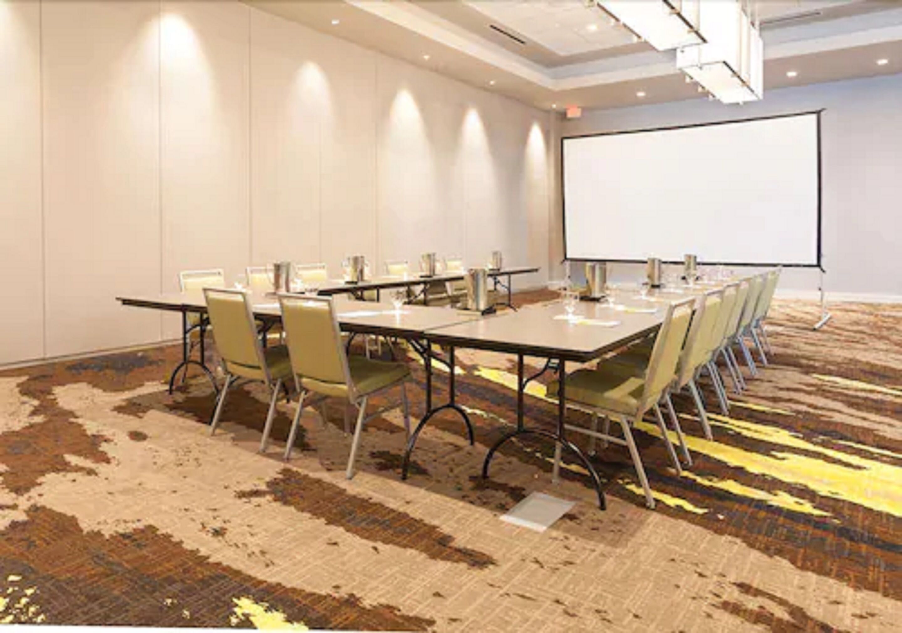 Un salón de conferencias con una mesa grande, sillas, una pantalla de proyector y suelo alfombrado.