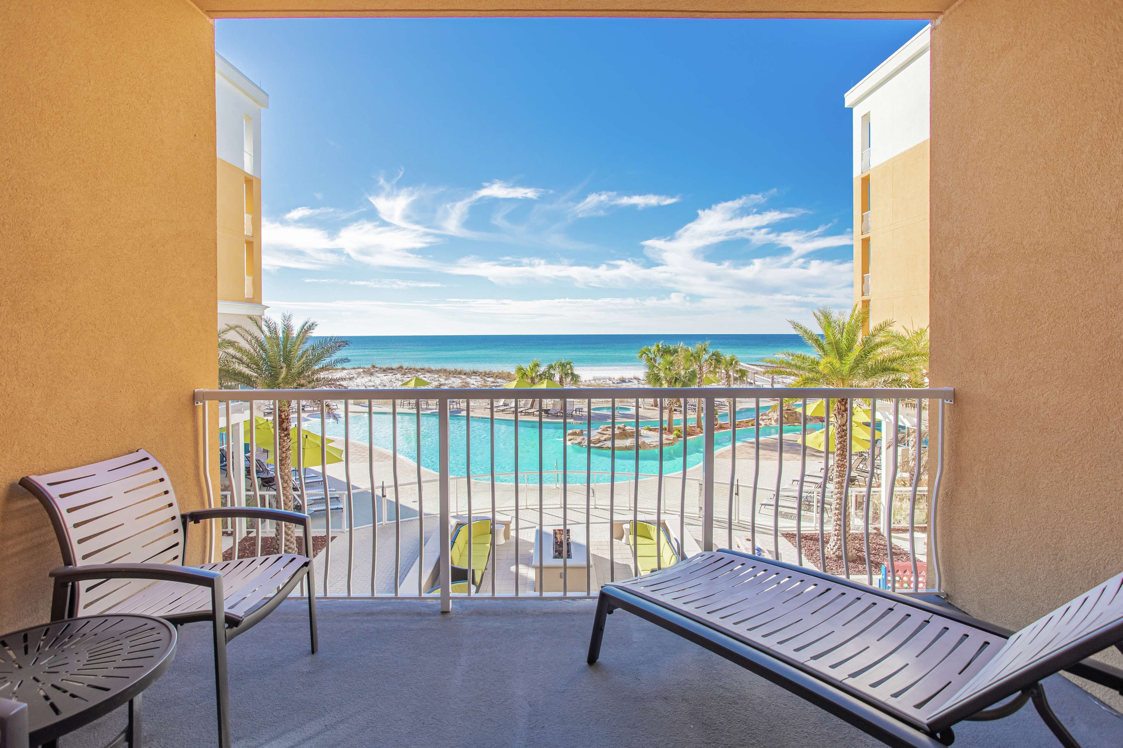 A balcony with two lounge chairs, a small table, and a view of a pool and beach.