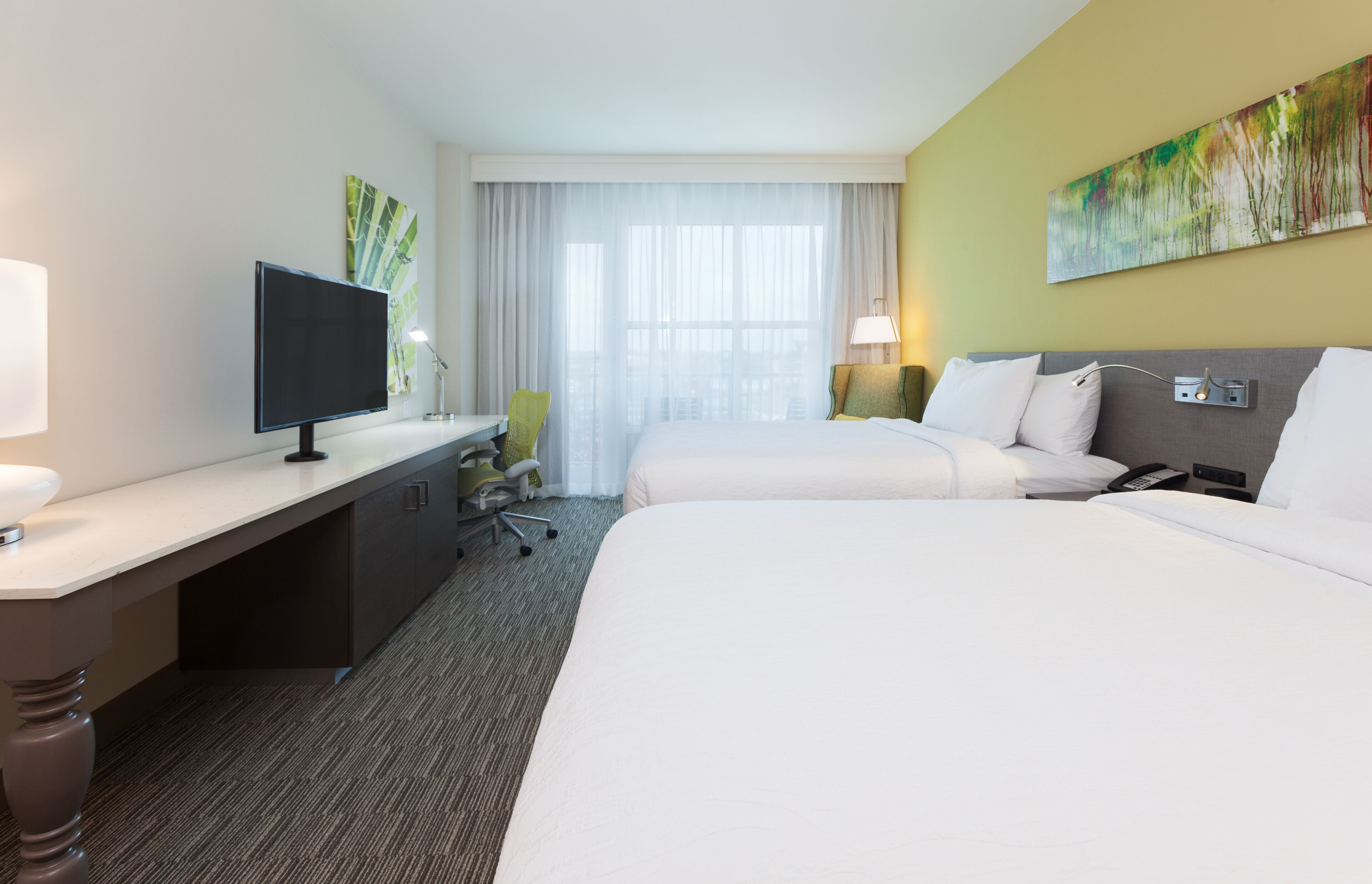 A hotel room with a desk, two beds, a TV, and a large window with sheer curtains.