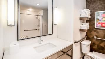 A bathroom with a white sink, mirror, and toilet. There are towels stacked on a shelf, and a shower area is visible in the background.