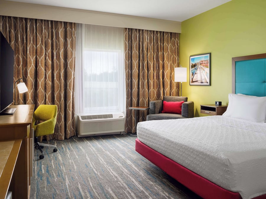 Ein Hotelzimmer mit einem großen Bett, einem Schreibtisch mit Stuhl, einem kleinen Tisch mit Lampe und einem Fenster mit Vorhängen.
