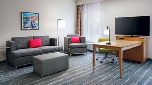 Een hotelkamer met een grijze bank, een klein fauteuil, een houten bureau, een televisie en een lijst met een afbeelding aan de muur.