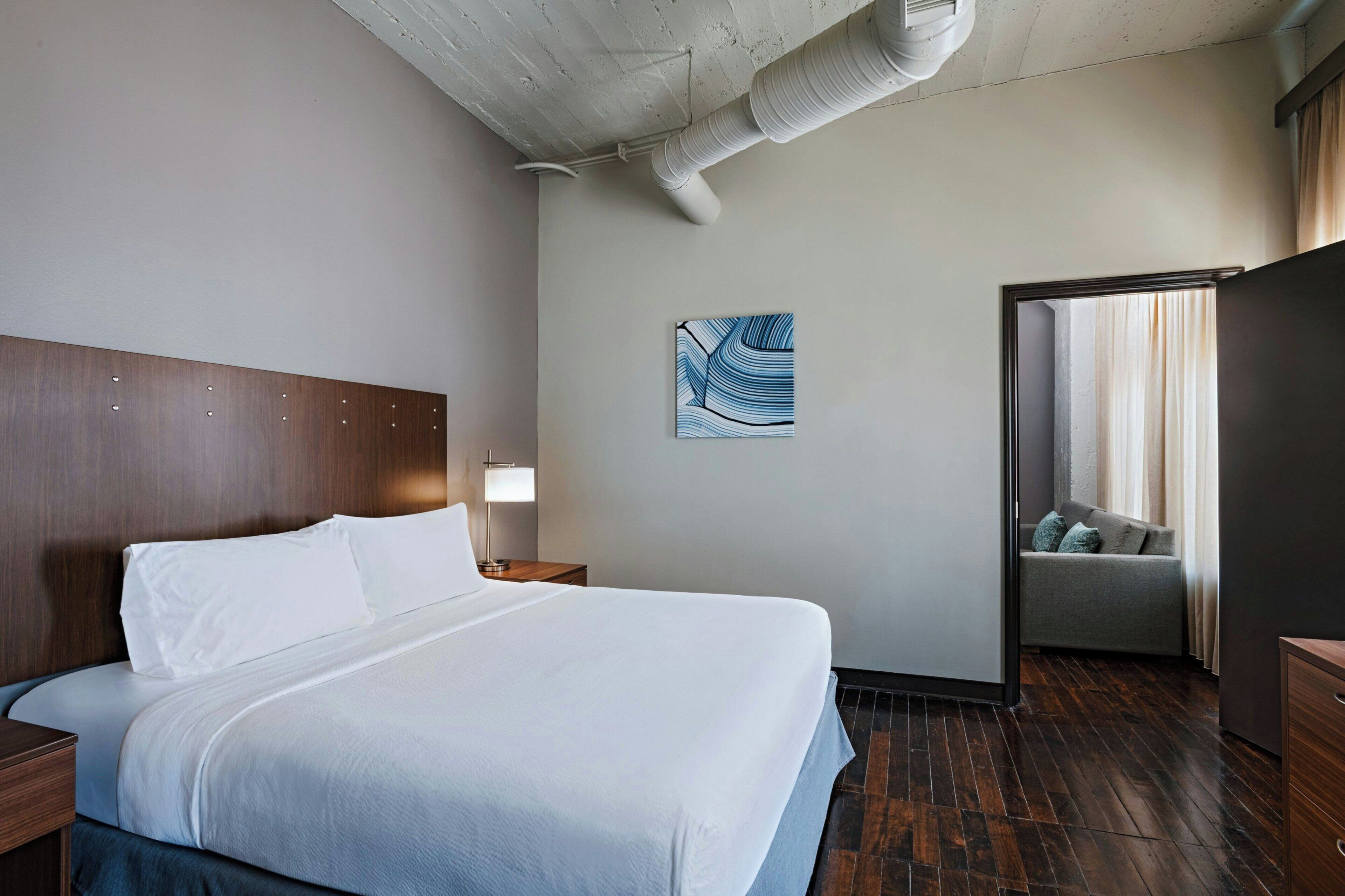 A hotel room with a large bed, a nightstand, a wall-mounted lamp, a painting, a sliding door to a seating area, and a wooden floor.