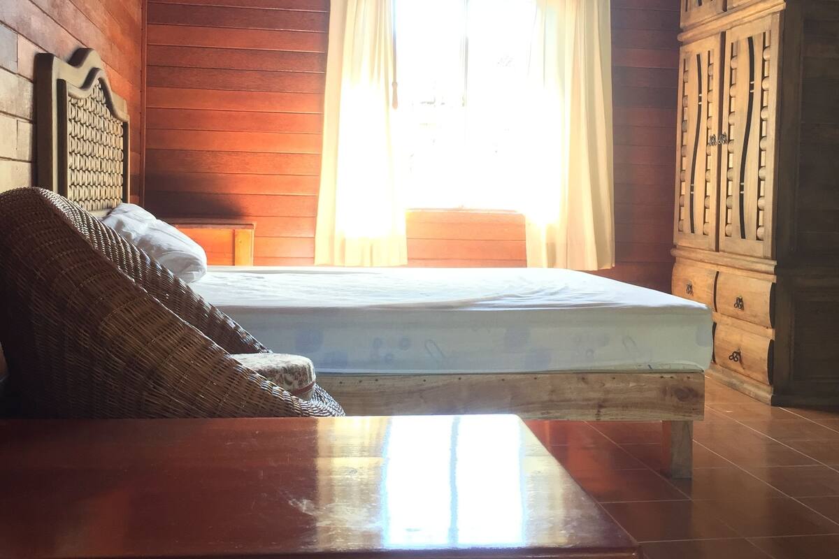 Interior de una cabaña de madera con escaleras, una ventana con cortinas y un sillón tejido.