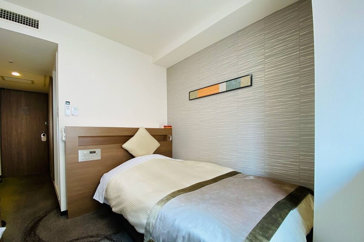 A hotel room with a single bed, a wooden headboard, a wall-mounted control panel, and a door leading to another room.