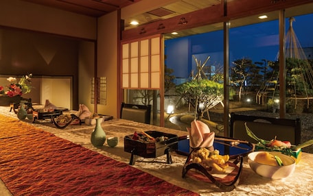A dining area with a long table, chairs, and a view of a garden at dusk.