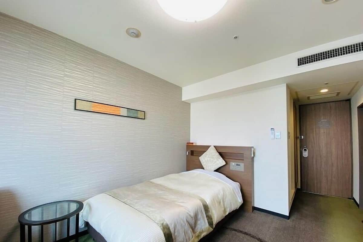 A hotel room with a single bed, a small round table, a wall-mounted light fixture, and a door leading to another room.