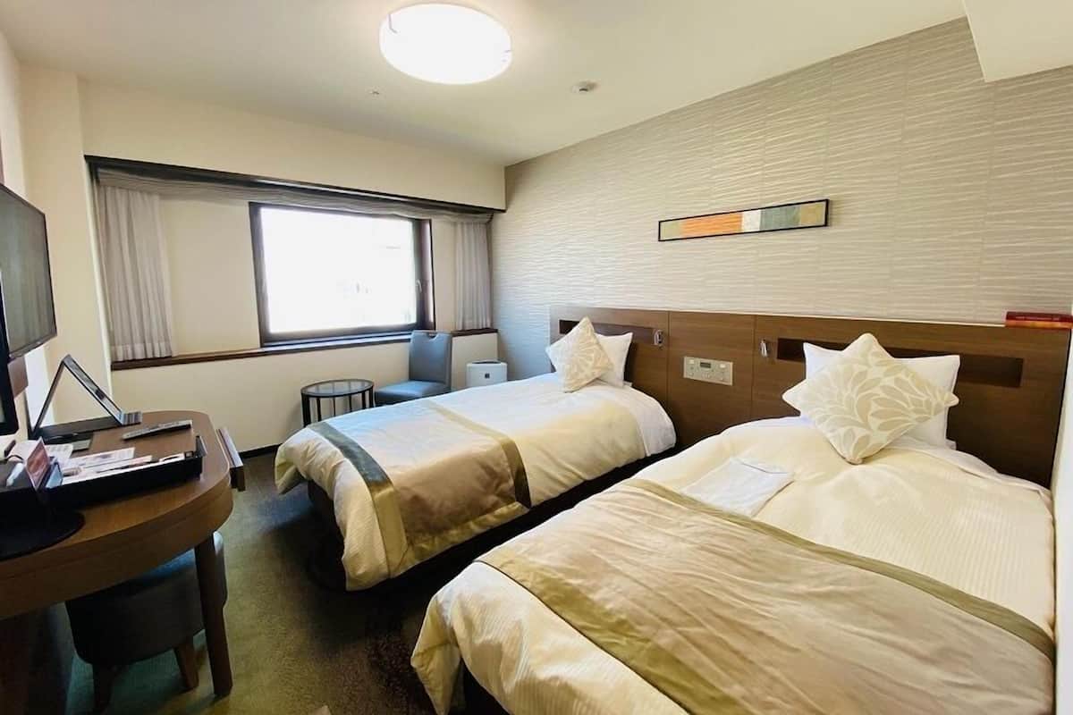 A hotel room with two beds, a desk with a laptop, a chair, and a window with curtains.