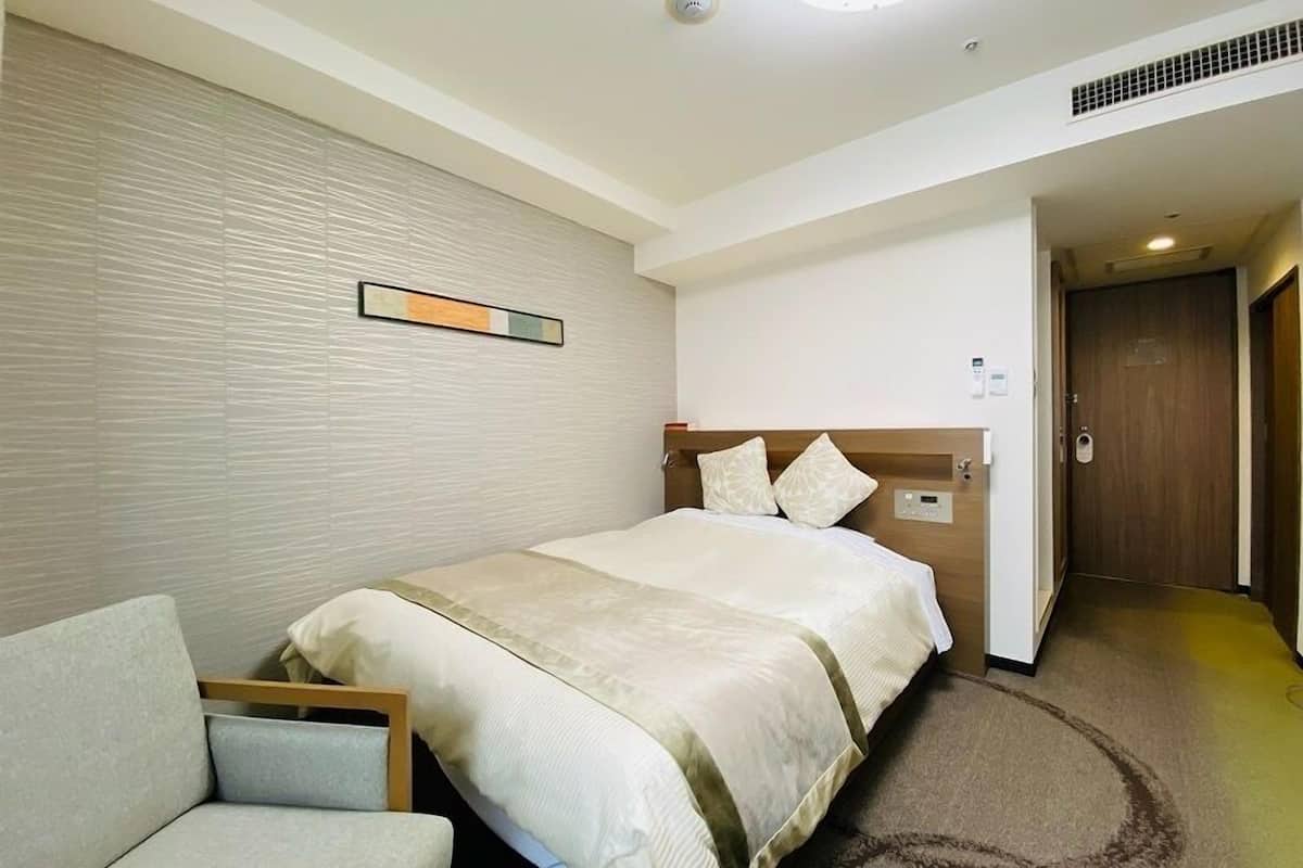 A hotel room with a bed, a chair, a wall-mounted control panel, and a door leading to another room.