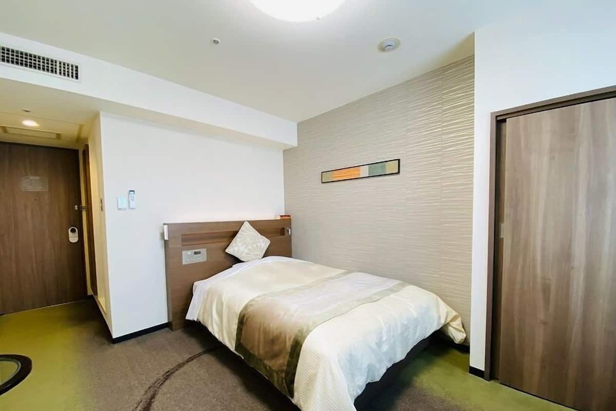A hotel room with a bed, a wooden door, a wall-mounted control panel, and a carpeted floor.