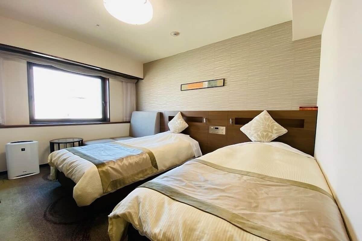A hotel room with two beds, a window, a wall clock, and a small table.