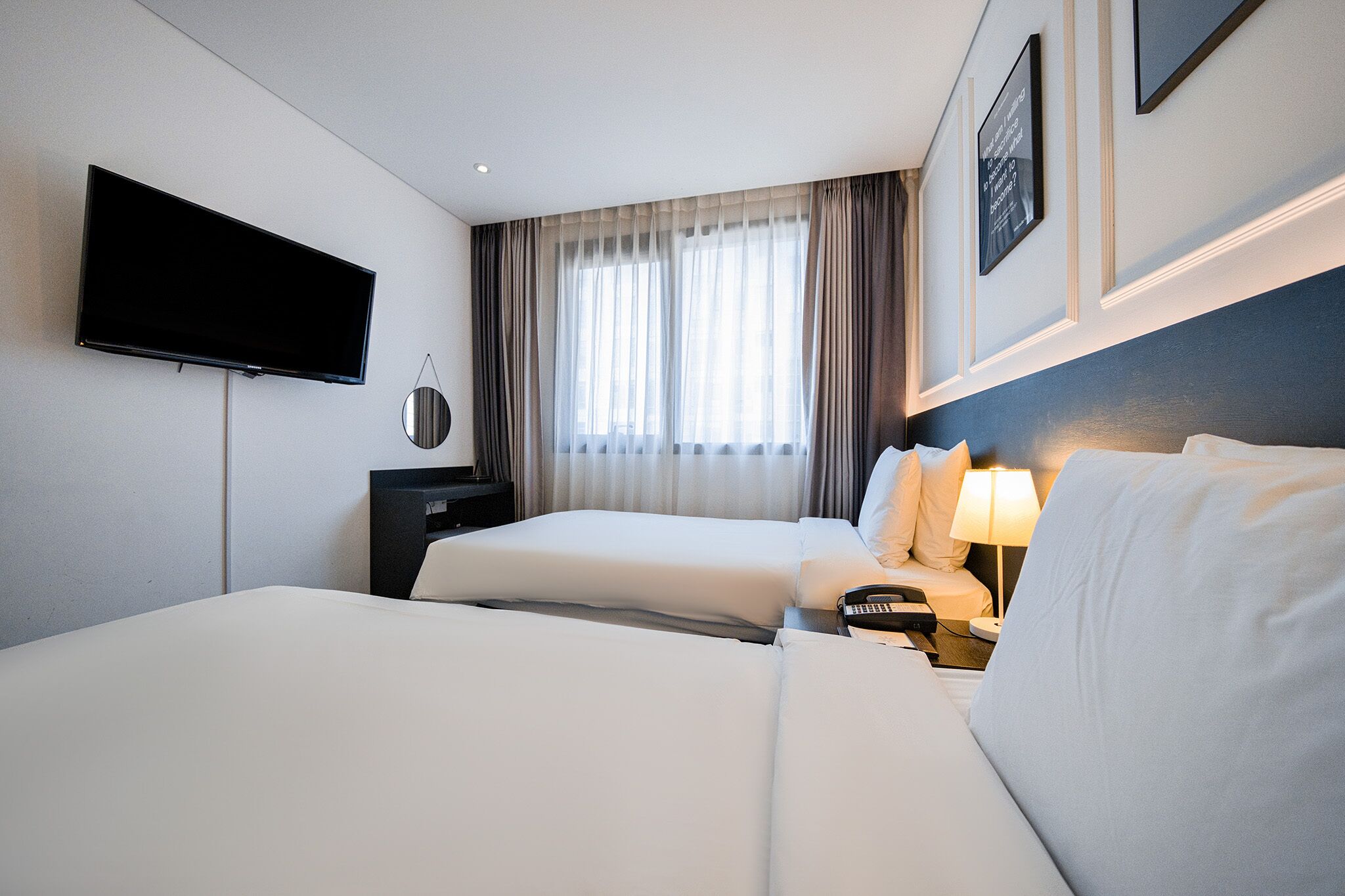 Une chambre d’hôtel avec deux lits, un téléviseur fixé au mur, une table de chevet avec une lampe et une fenêtre avec des rideaux.