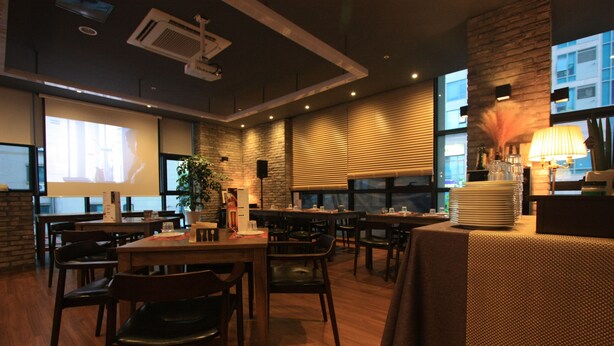 Een restaurant met meerdere tafels en stoelen, een projectiescherm en een groot raam met zonwering.