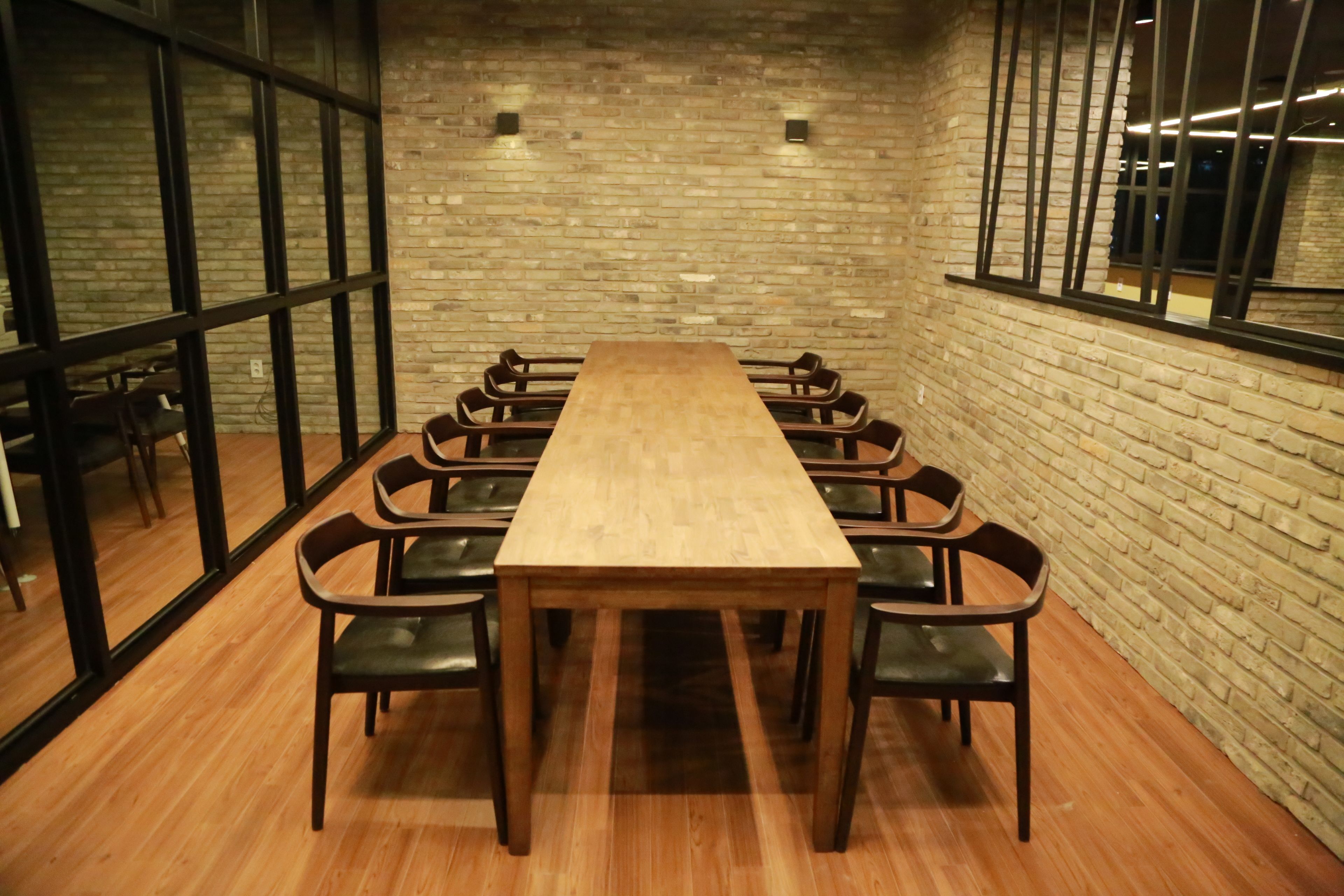 A conference room with a long wooden table and black chairs, brick walls, and large windows.