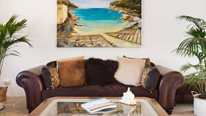 Een woonkamer met een bruine leren bank, een glazen salontafel met boeken en een decoratie, en een groot schilderij van een kustlandschap aan de muur.