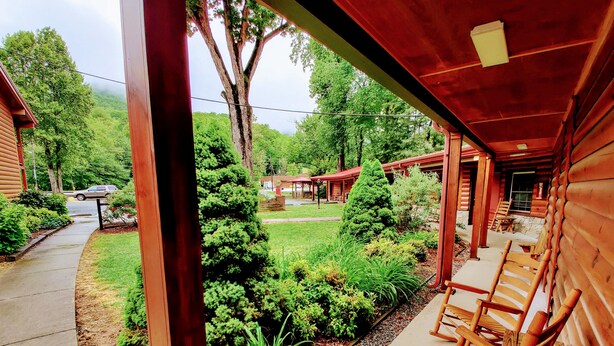 A wooden cabin with a porch, a small garden, and a view of a forested area.