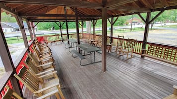 A covered picnic area with wooden benches and picnic tables, a wooden deck, and a view of a road and greenery.