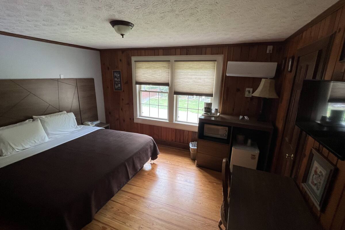 A bedroom with a bed, wooden walls, a window with blinds, a desk with a microwave, and a television.