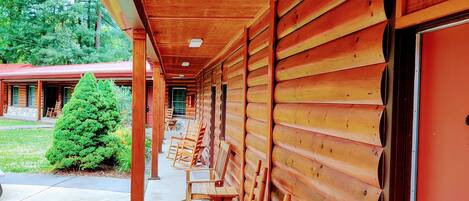 Une rangée de fauteuils à bascule en bois se trouve sous un porche abrité, dont la paroi extérieure est également en bois et la porte est peinte en rouge.