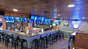 A bar with multiple screens displaying sports events, a counter with bar tools, and several black bar stools.