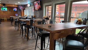 A restaurant interior with wooden tables and black chairs, a bar area, and multiple TVs on the walls.