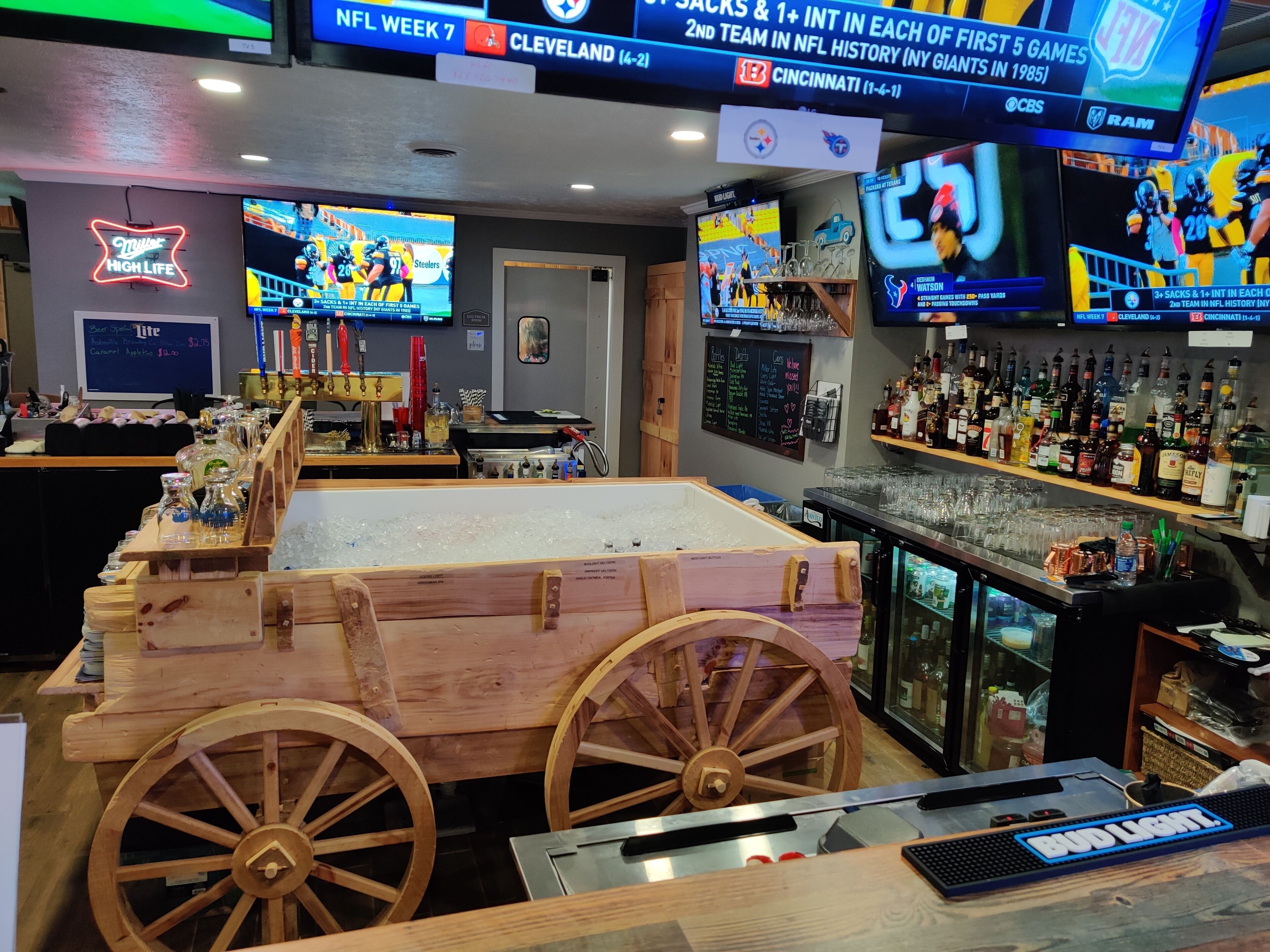 A bar area with multiple TVs displaying sports games, a wooden cart, and a variety of bottles and glasses on shelves.